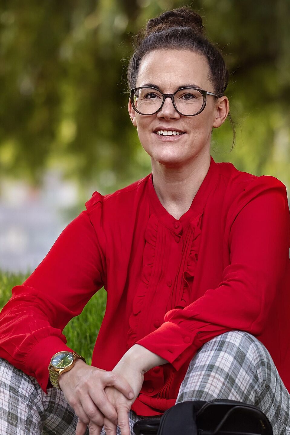 Katja Diehl Eine Frau sitzt im Gras, trägt eine rote Bluse und lächelt in die Kamera